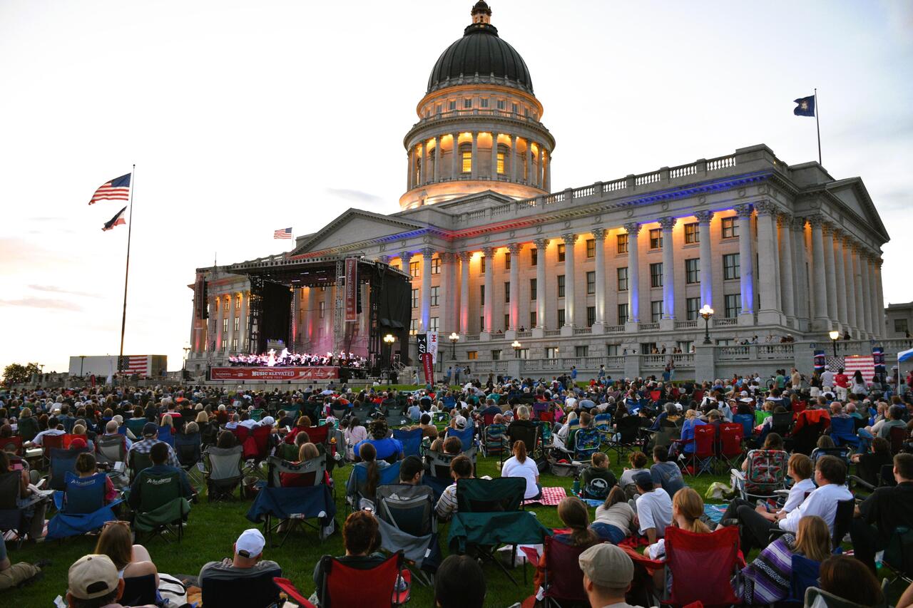 Large crowd watches an outdoor concert on the lawn in front of a government building at dusk.