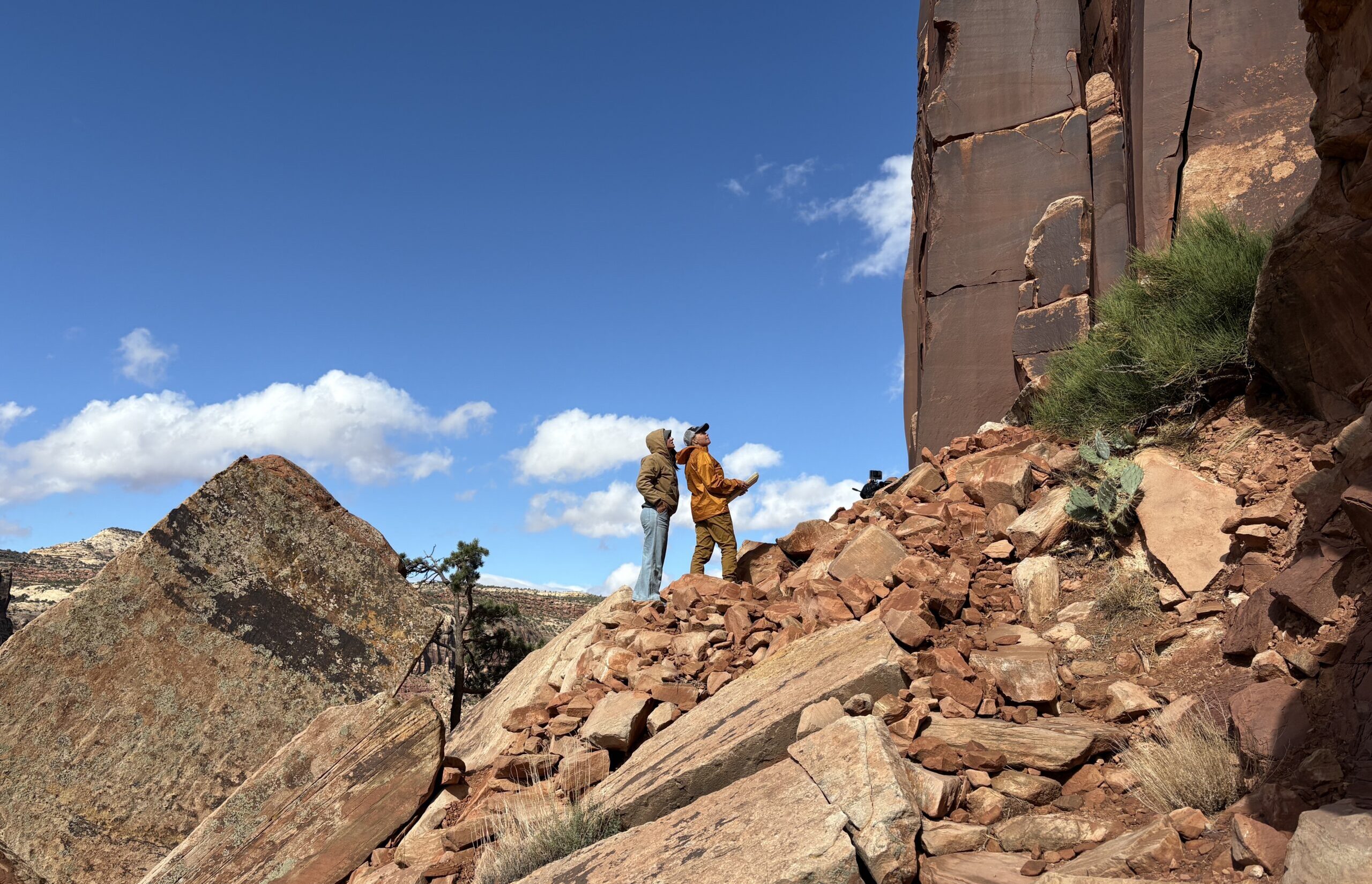 Two people stand on a rocky slope, looking up at a tall cliff face.