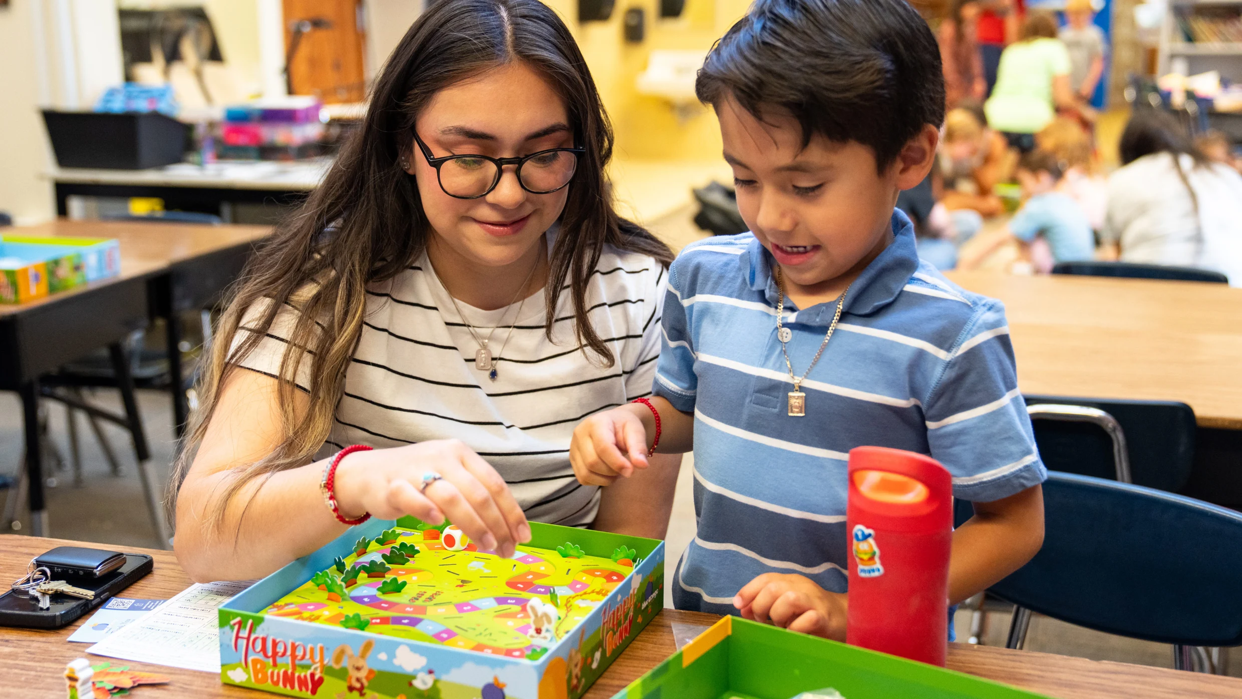 A woman and a young boy develop math skills while playing a Happy Bunny board game at a desk.